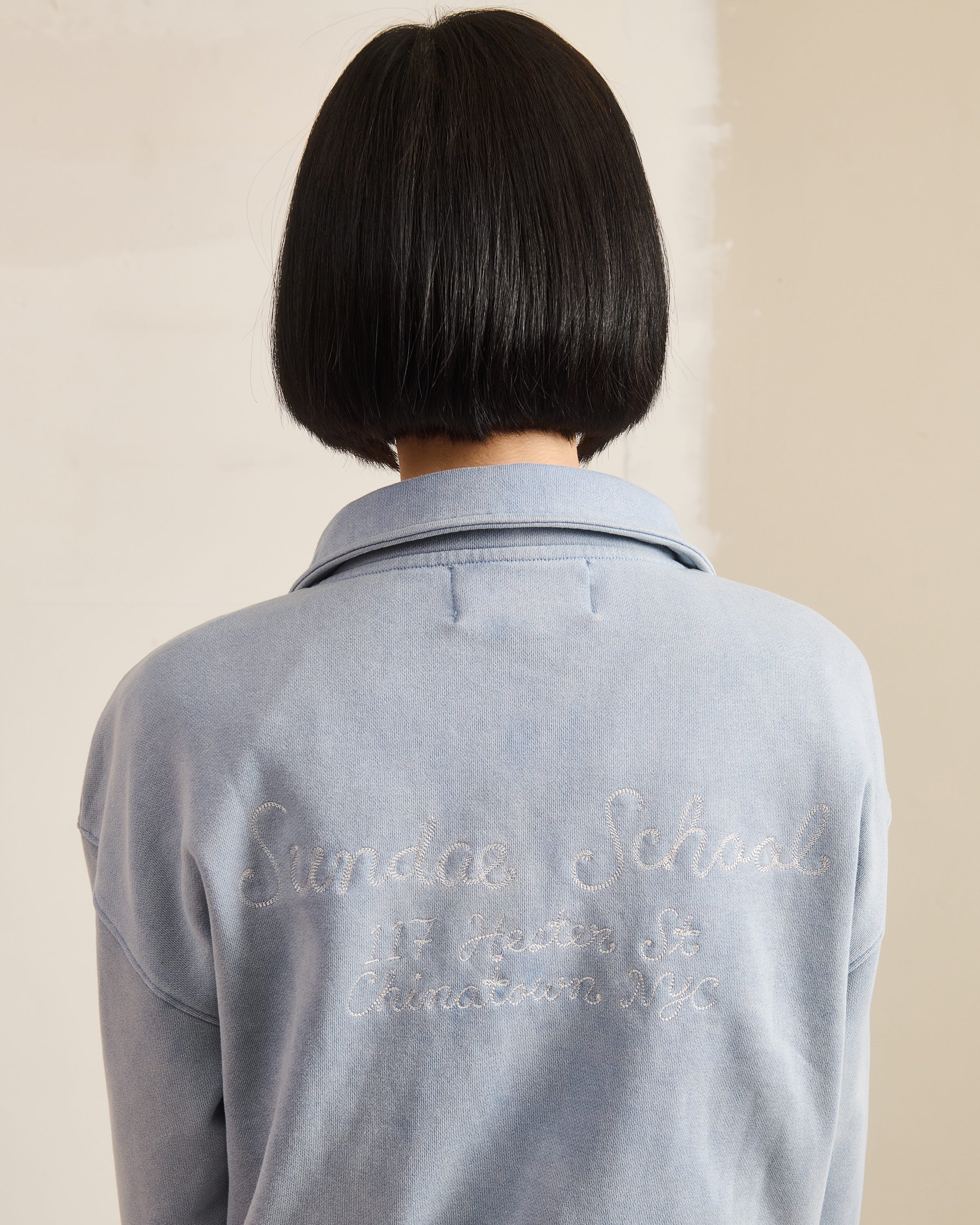 A person with a straight black bob is shown from behind, wearing the Sundae School SKY BLUE HESTER QUARTER ZIP, featuring cursive embroidery reading 
