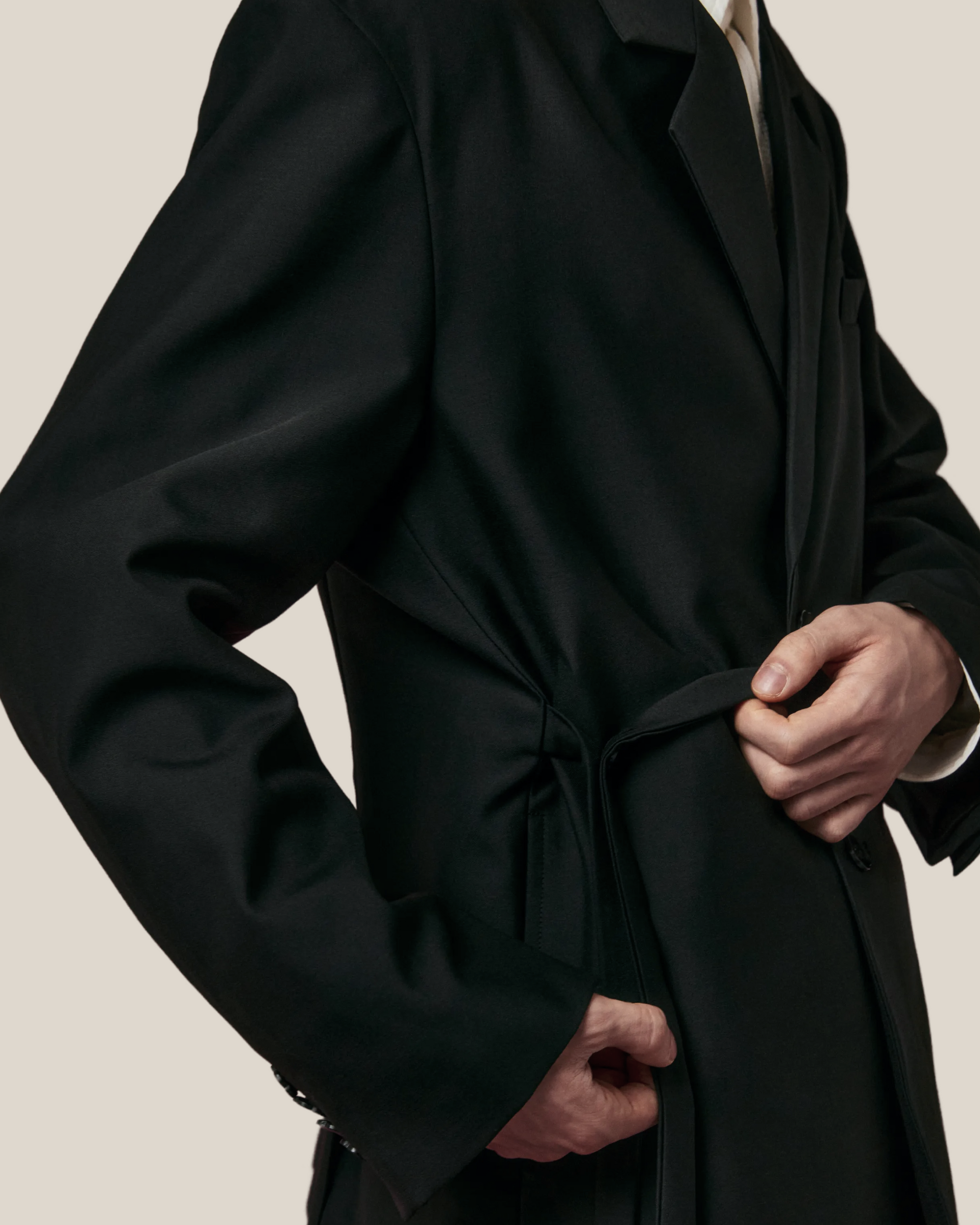 Side view of model wearing tailored double duty tie single-breasted blazer with notch lapels and waist tie gorum detail that can be hidden or cinched.