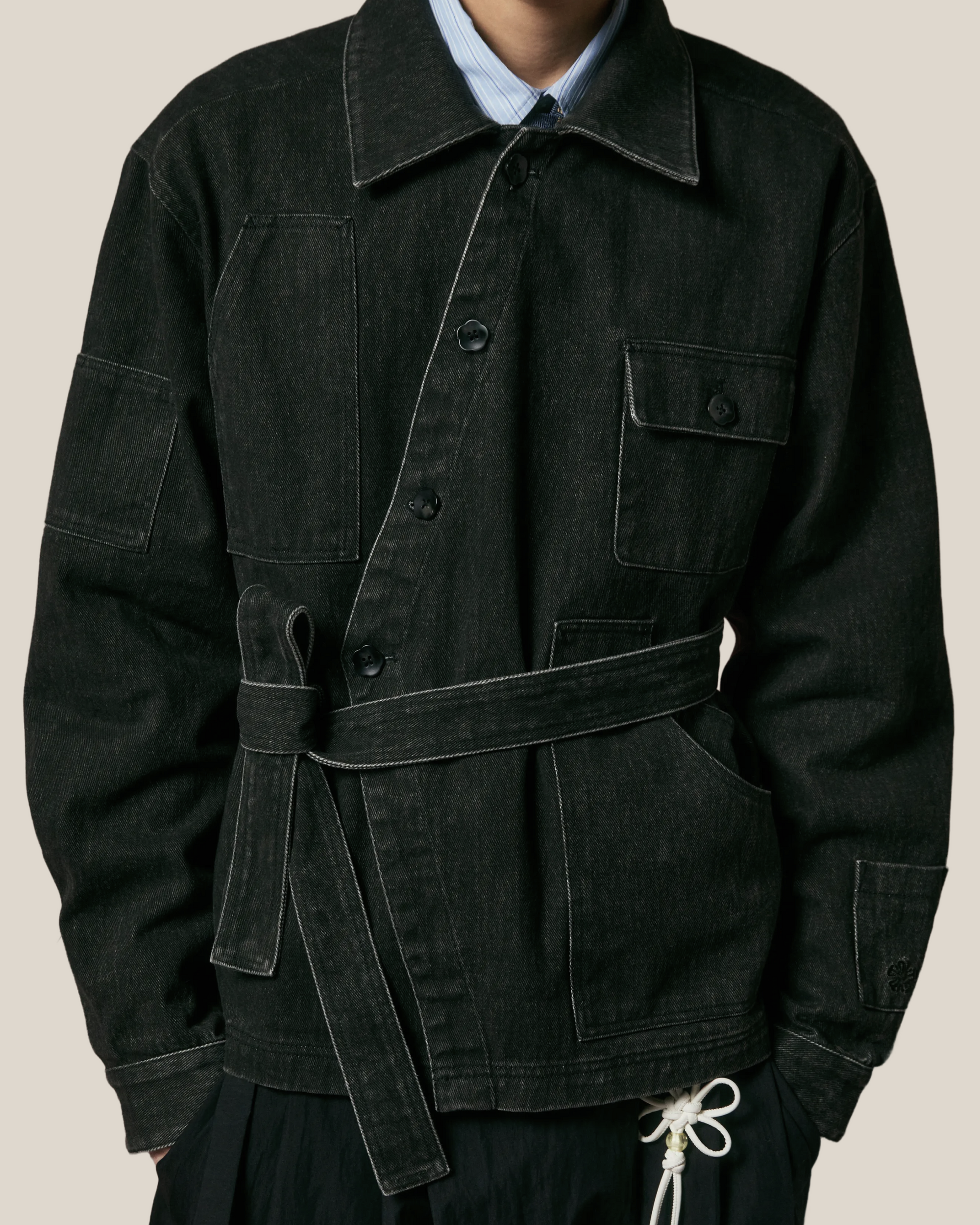 Model wearing black denim belted asymmetric work jacket with chest flap pocket, and contrast white stitching
