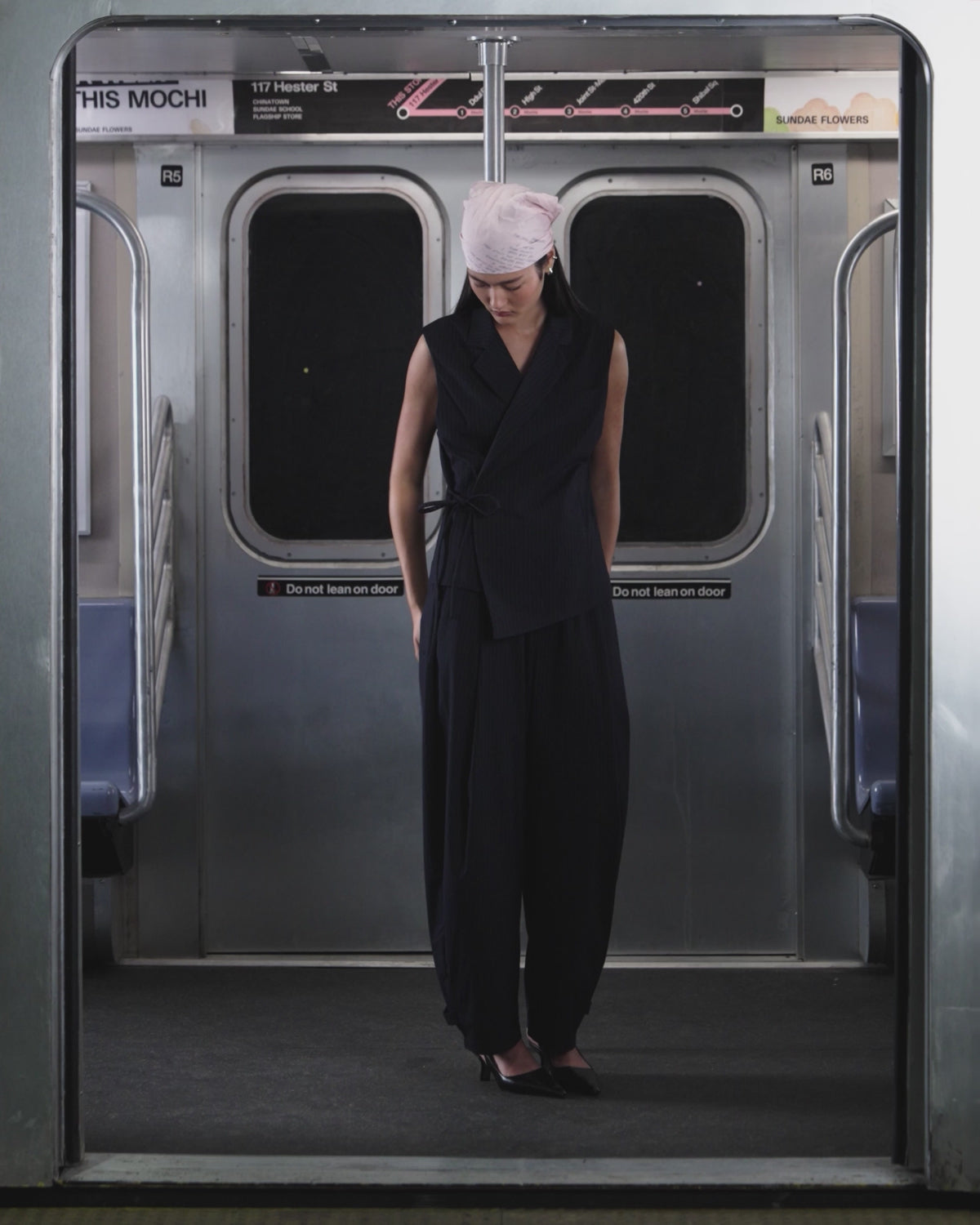 A person wearing the SABRINA VEST and KIKI DRAWSTRING SLACKS by Sundae School stands in a subway car, with “117 Hester St” and a subway map in the background.
