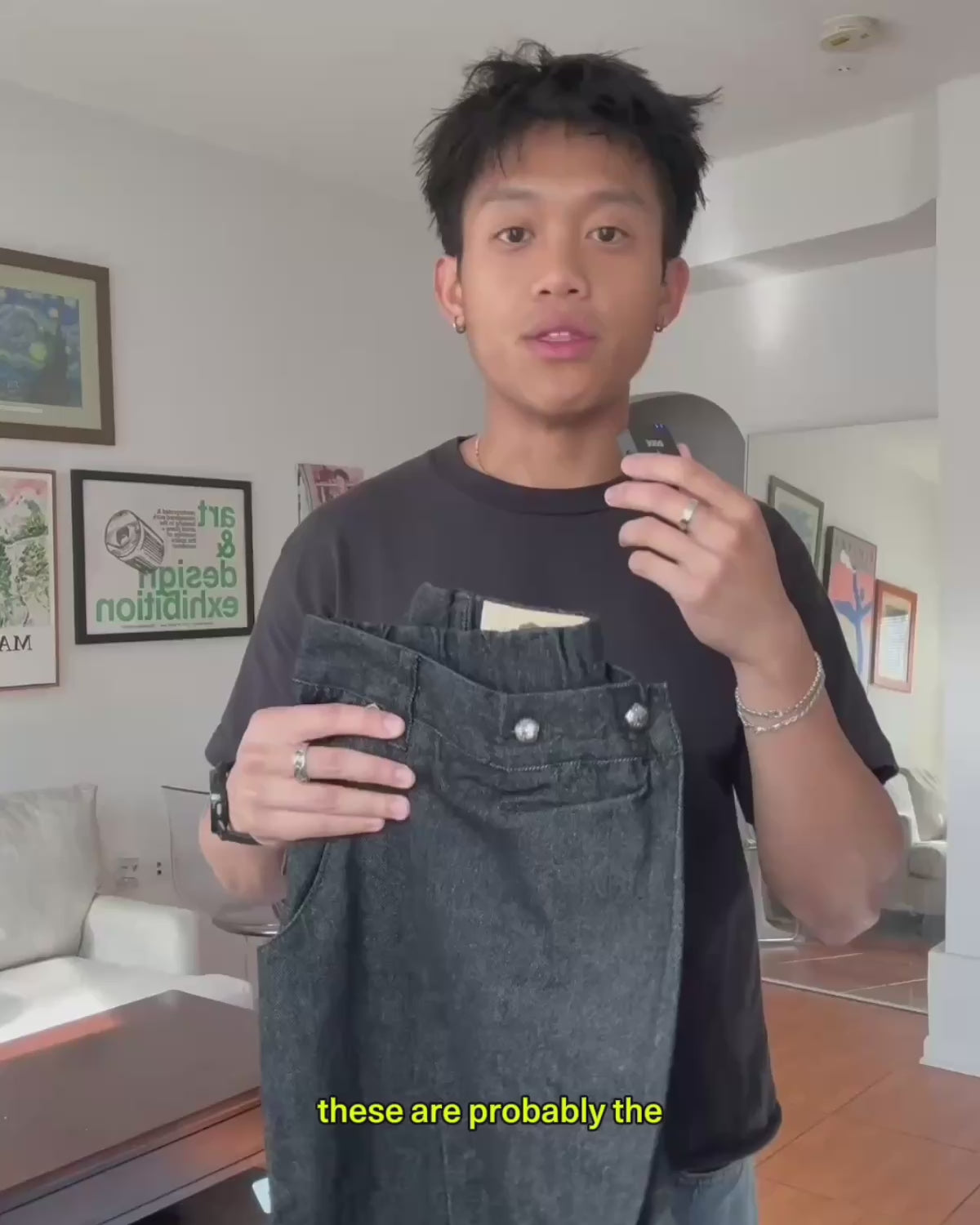 A product review and styling video of Sundae School's Black Hanbok Denim Pants. The presenter holds up the dark denim jeans to highlight key features—such as the asymmetrical adjustable waistband, flower-shaped metal buttons, back branded patch, and adjustable ankle cuffs—before showcasing a complete outfit wearing the pants.