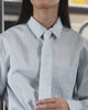 A person with long, straight dark hair wears the Sundae School NECKTIE SHIRT and FLARE SKIRT PANTS, a relaxed light blue shirt with a stash pocket, standing back to the camera inside a subway at 117 Hester St. A route map is visible above the doors.