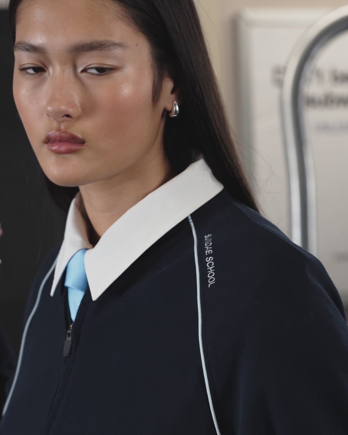 A woman with long dark hair and sunglasses wears the Sundae School NAVY OFFICE QUARTER ZIP, light blue tie, and dark shorts as she stands by subway doors beneath a map and a “117 Hester St” sign.