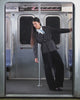 A woman stands in a subway car wearing the BLACK FLOWER NORIGAE LINEN TROUSERS from Sundae School, styled with a matching black suit and a white collared shirt. She faces forward, holding onto the metal rails and leaning back, between empty seats, with subway maps and signs visible behind her.