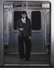 A woman with long, straight hair and a neutral expression stands in a subway car, wearing the Sundae School COLLARLESS TIE BLAZER with a light blue scarf and vegan leather belt. Behind her are a subway map and the 117 Hester St sign.