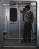 A person with curly hair, seen from behind in a Sundae School KHAKI OFFICE QUARTER ZIP, stands inside a subway train facing closed doors. Above the doors are a subway map and a sign that reads 117 Hester St.