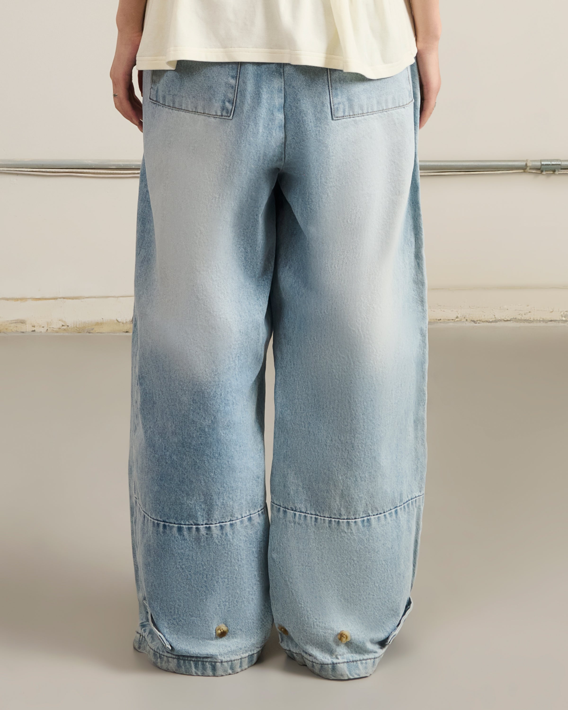 A person in Sundae School's SKY BLUE HANBOK DENIM PANTS with wide legs and large back pockets, shown from behind, stands on a smooth light-gray floor.