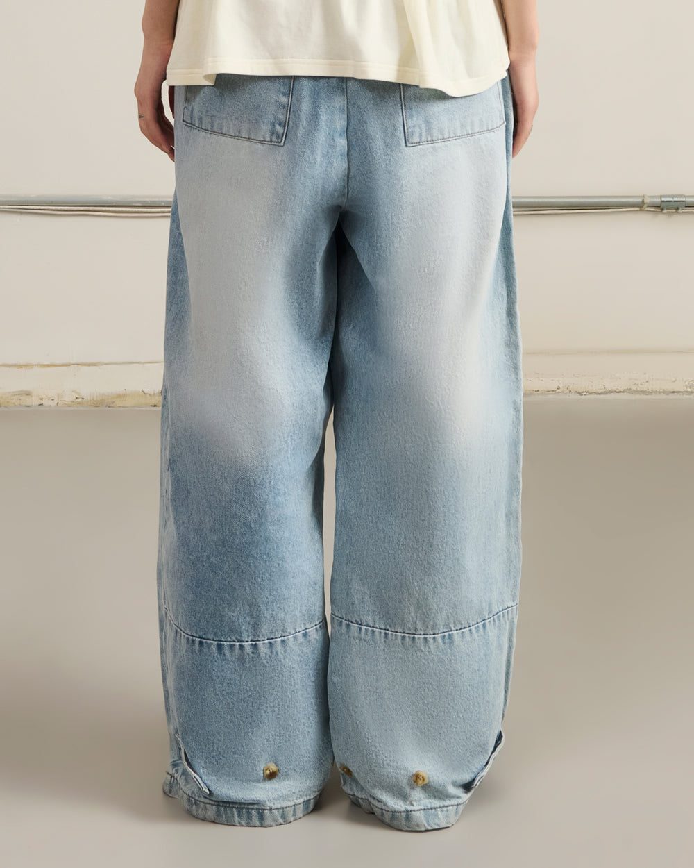 A person in Sundae School's SKY BLUE HANBOK DENIM PANTS with wide legs and large back pockets, shown from behind, stands on a smooth light-gray floor.