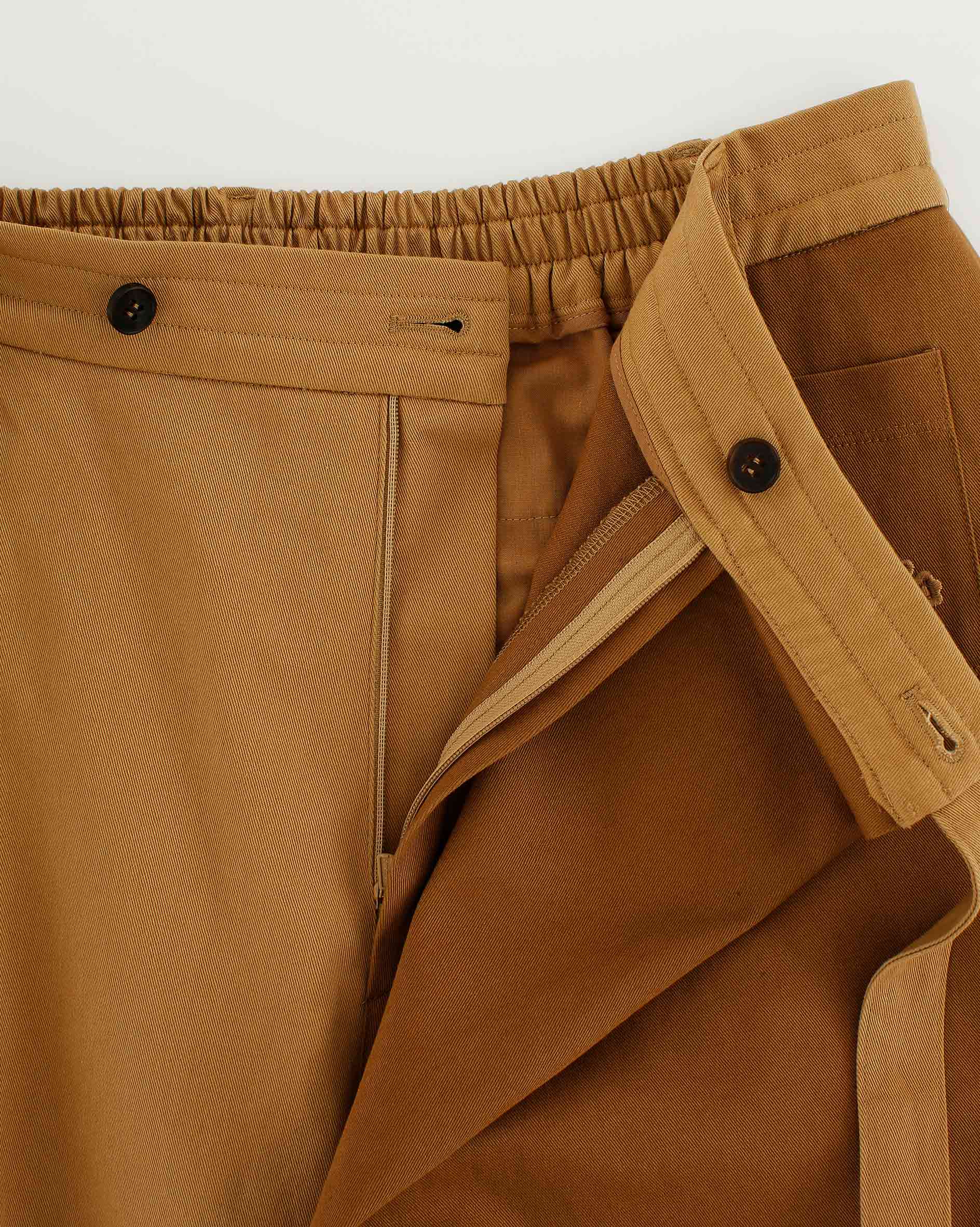 Close-up of Sundae School's Tan Pleated Hanbok Cargo Pants, inspired by Korean fashion, showcasing a tan and brown design with elastic waistband, button closures, a partially unzipped fly, and detailed stitching.