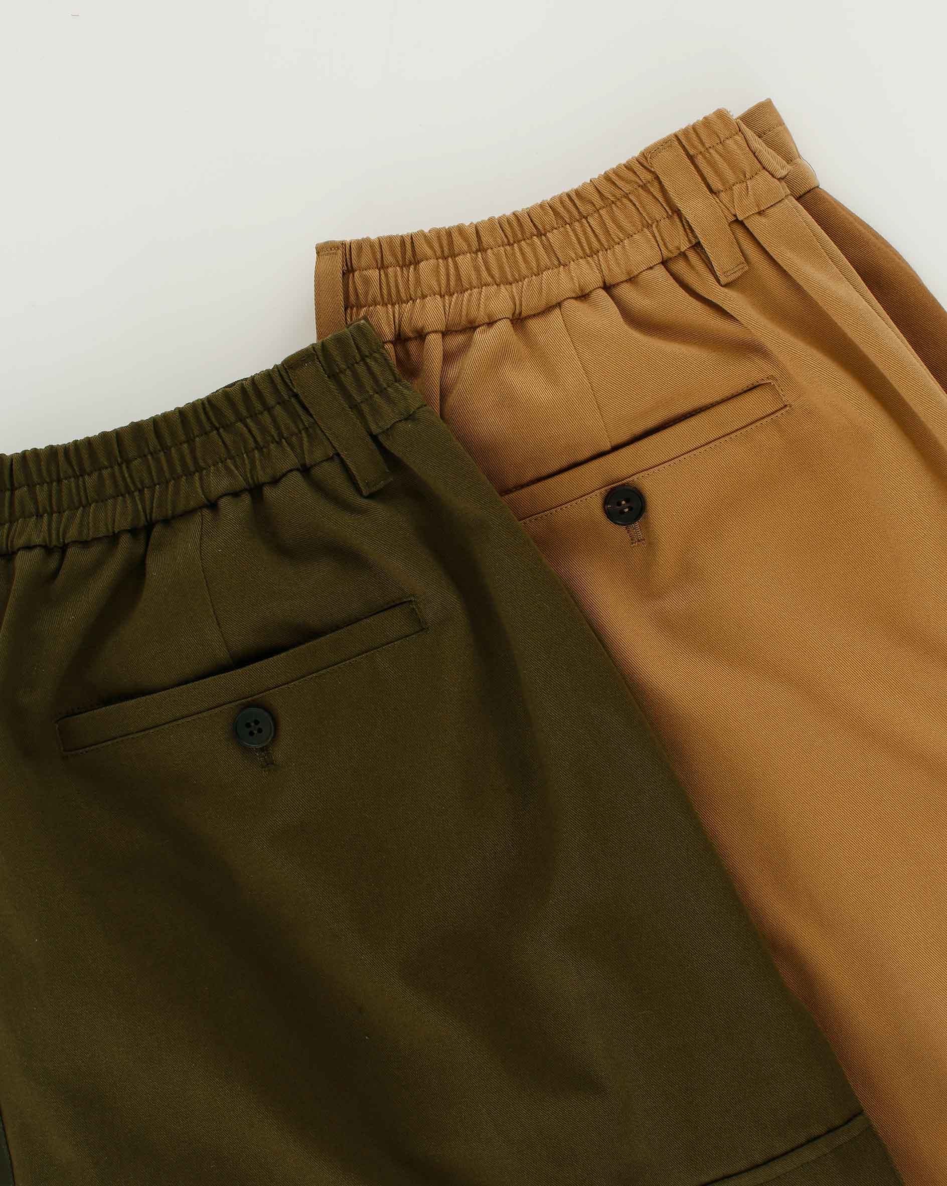 Two pairs of Sundae School Tan Pleated Hanbok Cargo Pants with elastic waistbands, in olive green and tan, are laid side by side. Each features a back pocket with a black button, showcasing modern Korean fashion.