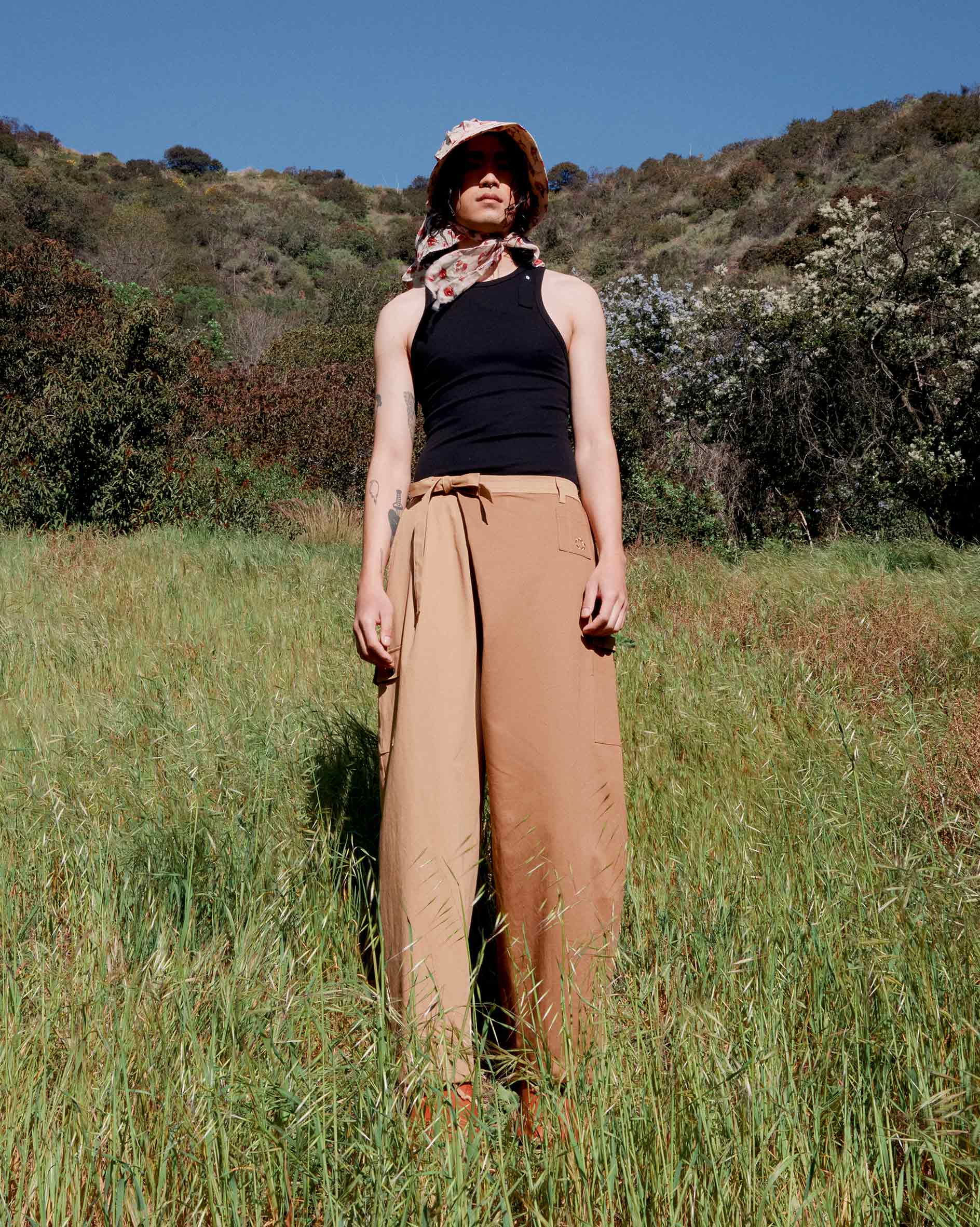 A person stands in a grassy field with hills and trees, wearing a black tank top, Sundae School Tan Pleated Hanbok Cargo Pants, and a floral sun hat with a scarf under a clear blue sky.