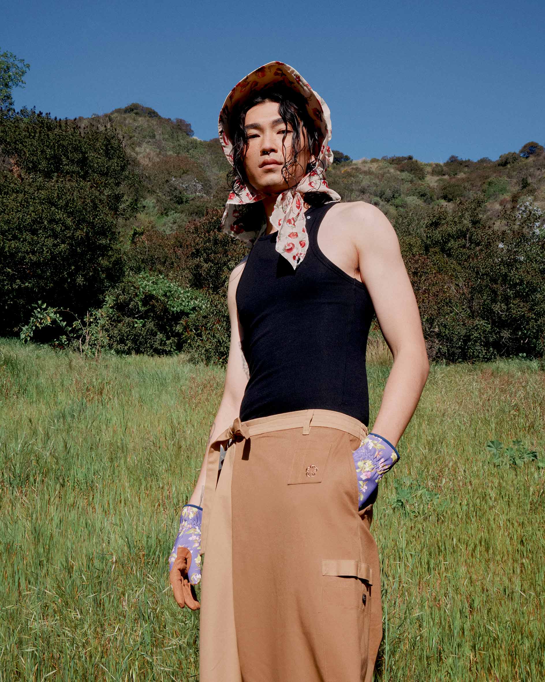 In a grassy field with green hills and blue sky, a person showcases modern Korean fashion in a black sleeveless top, Tan Pleated Hanbok Cargo Pants by Sundae School, purple floral gloves, and a patterned headscarf.